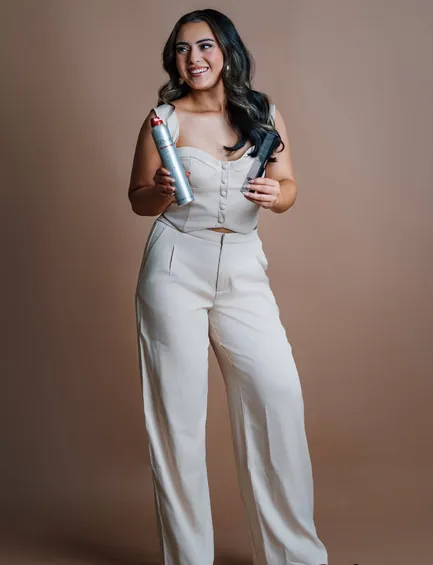 Nevaeh Perez in a cream corset top and wide-leg trousers, holding hairspray and a comb, with curling irons at her feet